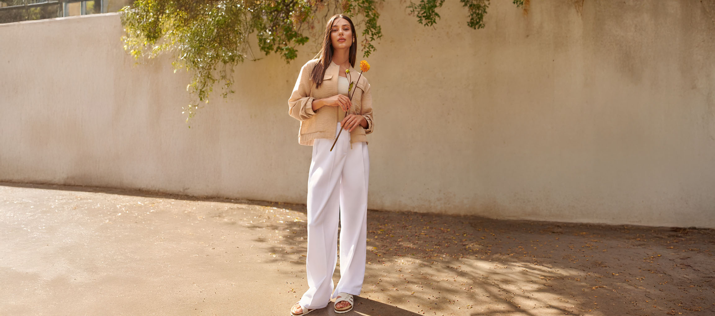 Nő bézs kabátban, fehér nadrágban és szandálban, sárga virágot tartva.