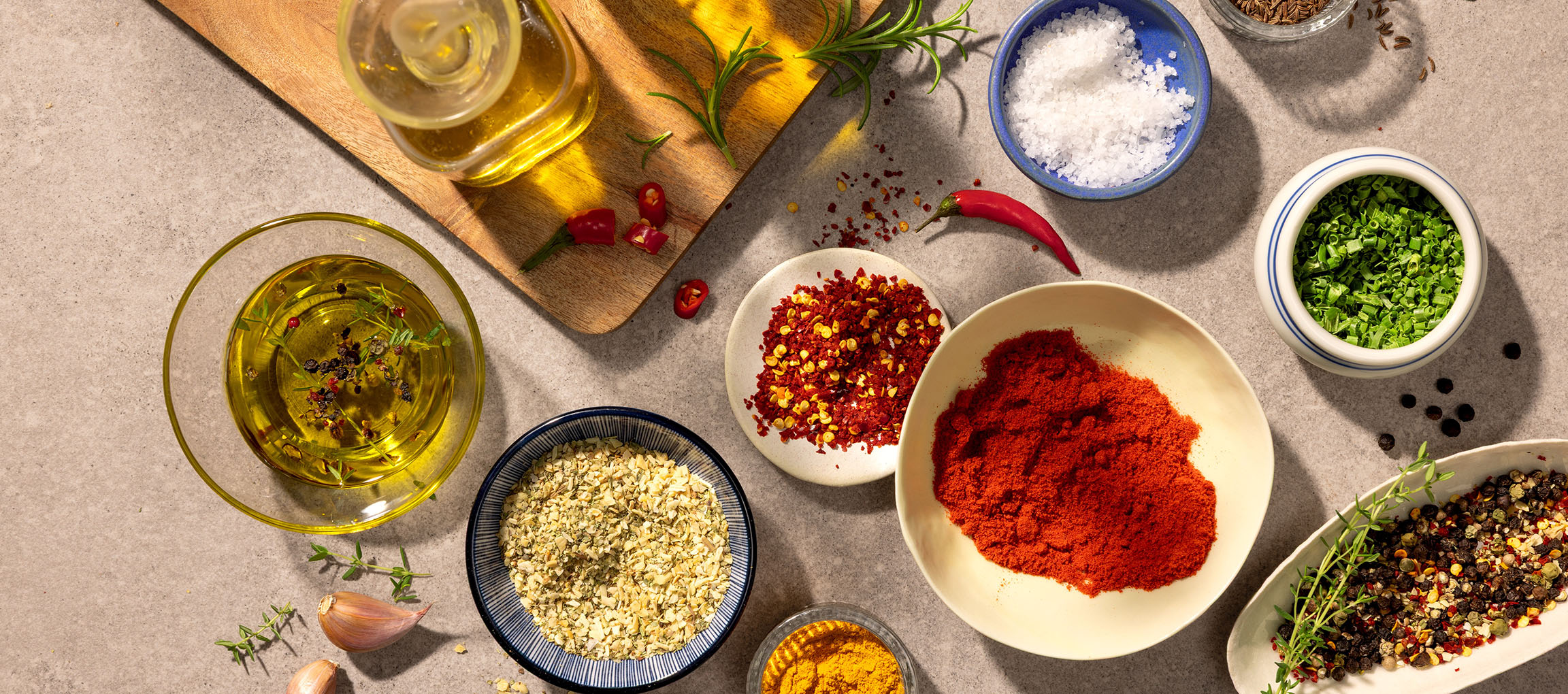 Különféle fűszerek és olajok, köztük paprika, chili, só, bors és olívaolaj, szürke háttéren.