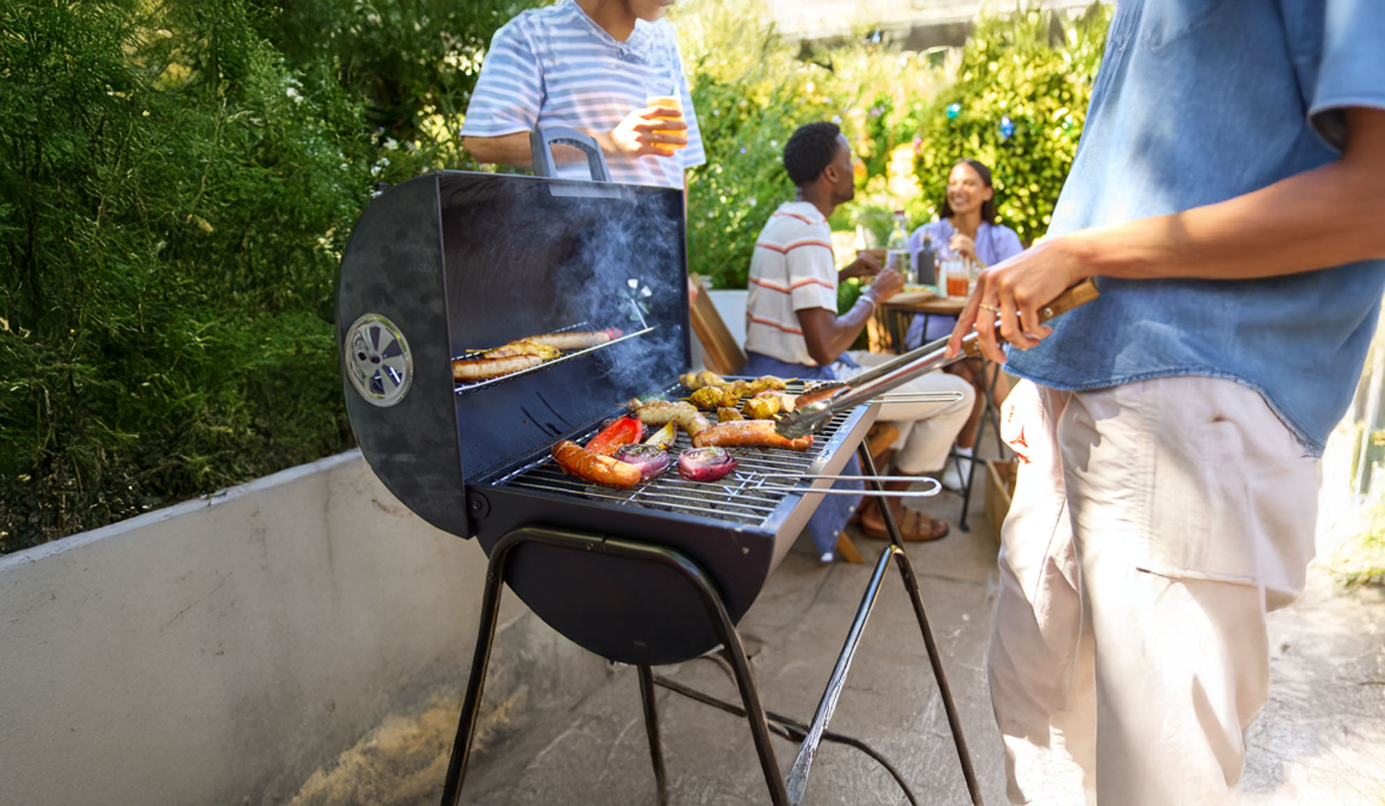 Barátok grilleznek kolbászt és zöldségeket a kertben.