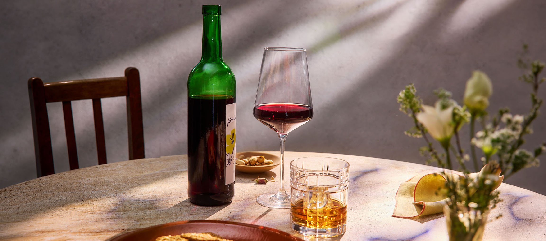 Egy üveg vörösbor, egy pohár bor és whisky az asztalon, székkel és virágokkal a háttérben.