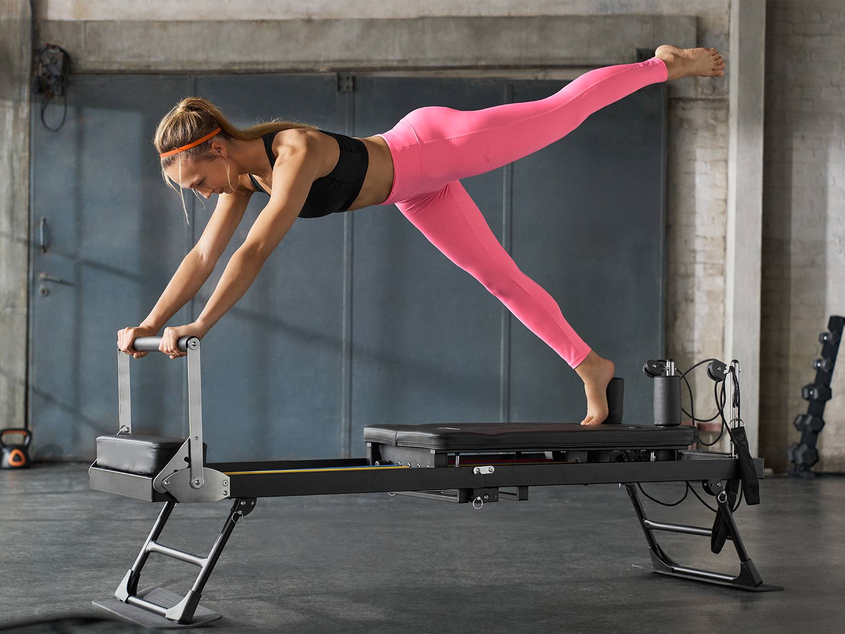 Nő rózsaszín leggingsben és fekete sportmelltartóban pilates reformeren edz.