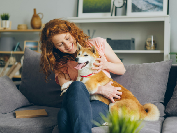 Egy mosolygós, vörös hajú nő a kanapén ülve öleli és simogatja Shiba Inu kutyáját.