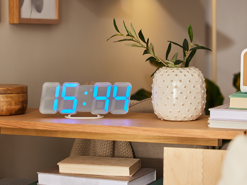 Modern LED digitális óra egy fa polcon, növényekkel és könyvekkel körülvéve.