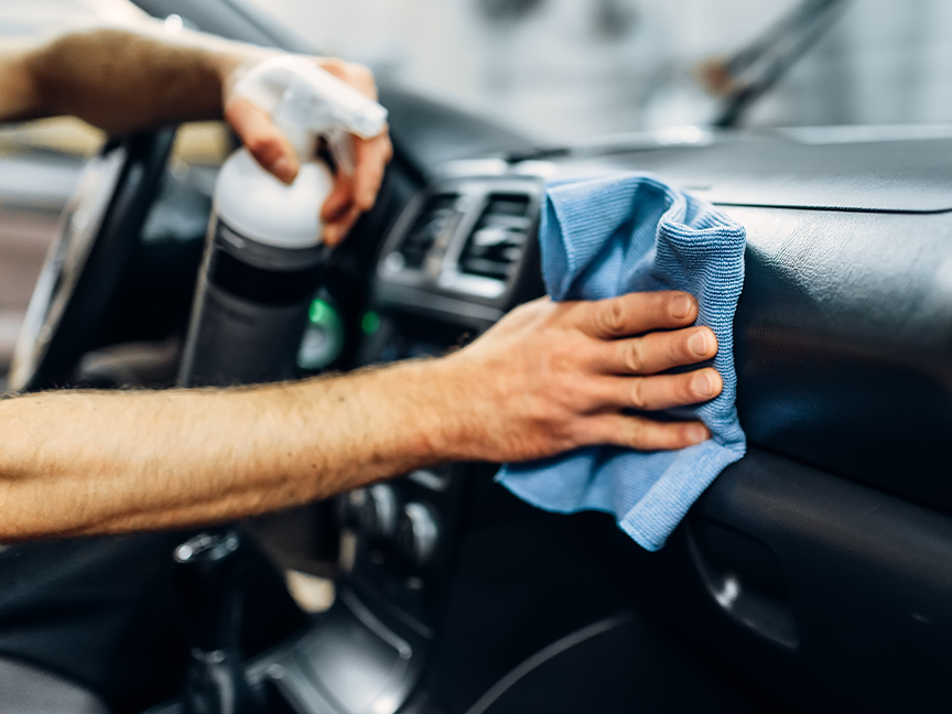Egy személy autó műszerfalát tisztítja egy spray-palackkal és egy mikroszálas kendővel.