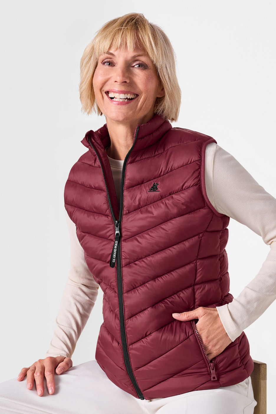 Női bordó steppelt mellény, bézs hosszú ujjú felsővel és fehér nadrággal.