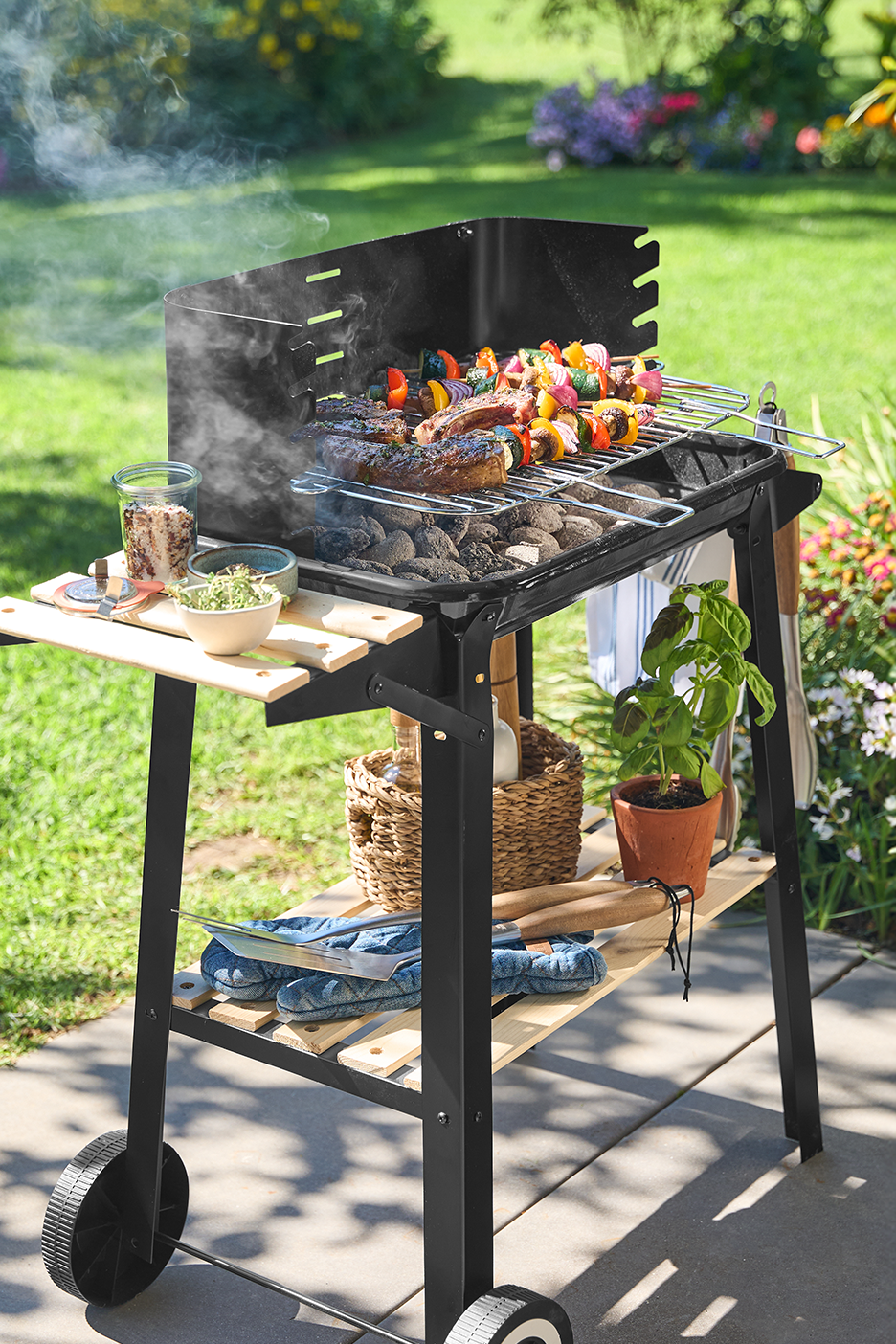 Füstölgő faszenes grill húsokkal és zöldségnyársakkal, kerti környezetben.