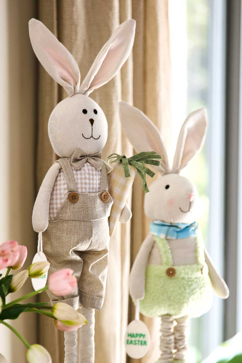 Két húsvéti nyuszi figura, az egyik kockás ingben és kantáros nadrágban, a másik zöld kantáros ruhában, mindkettőn 'Happy Easter' feliratú címke.