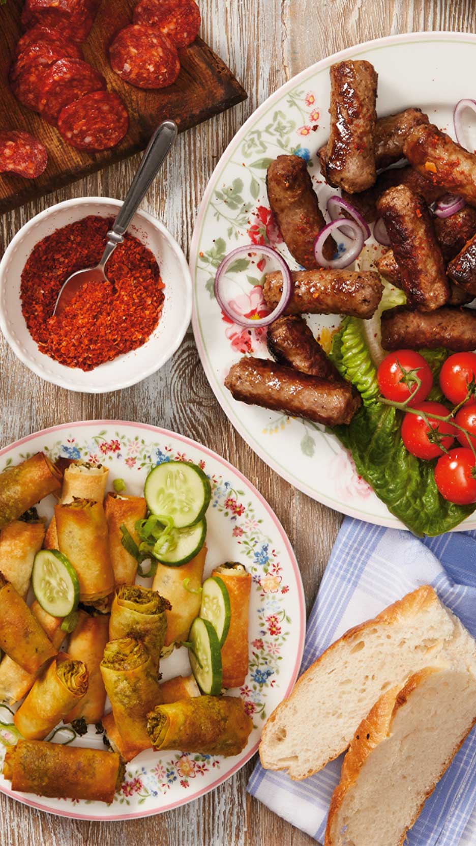 Grillezett kolbászok, tavaszi tekercsek, szalámi és fűszeres paprika egy asztalon.