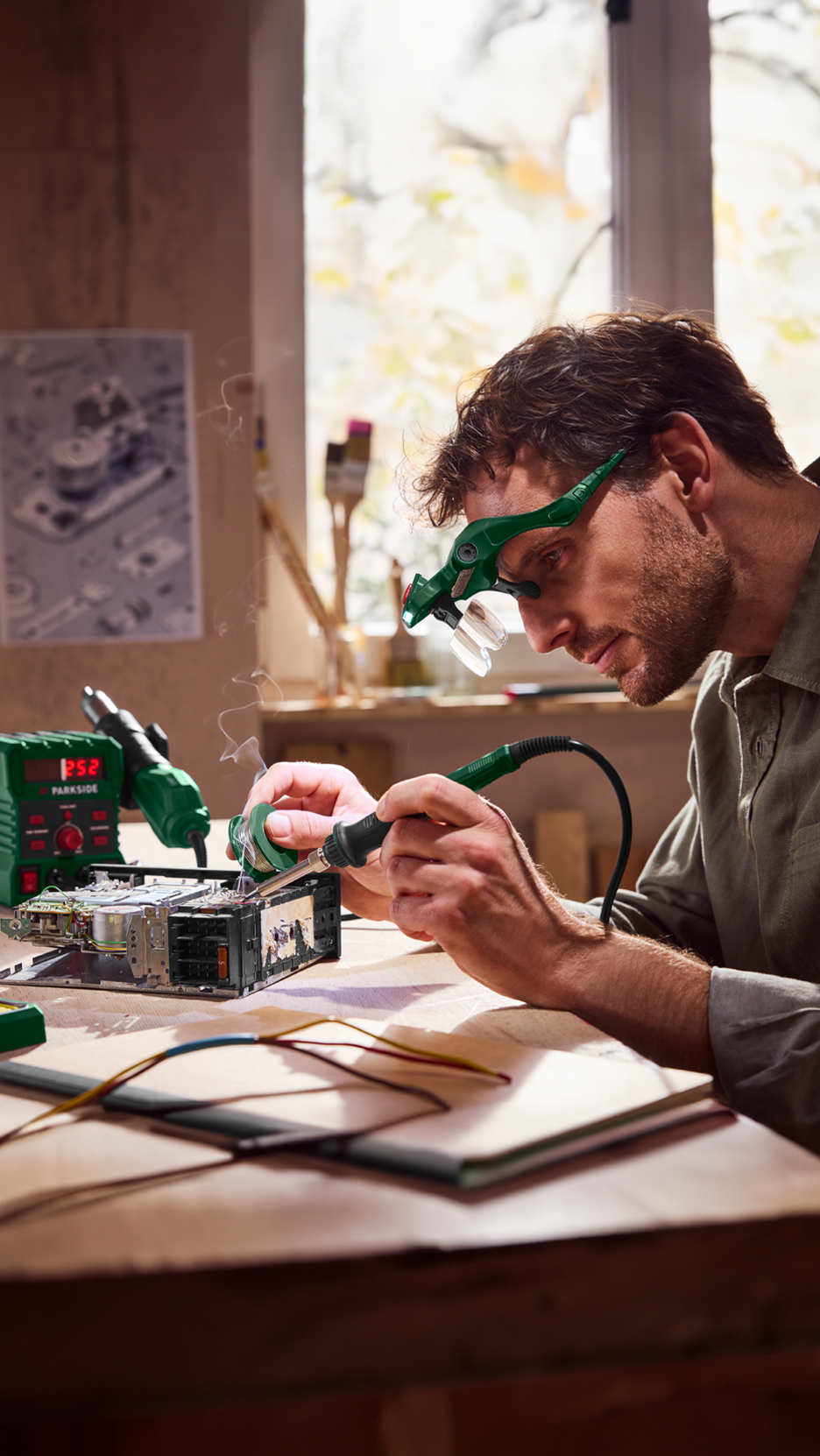 Férfi forrasztópákával és forrasztóónnal dolgozik egy elektronikai eszközön, nagyítóval a fején.