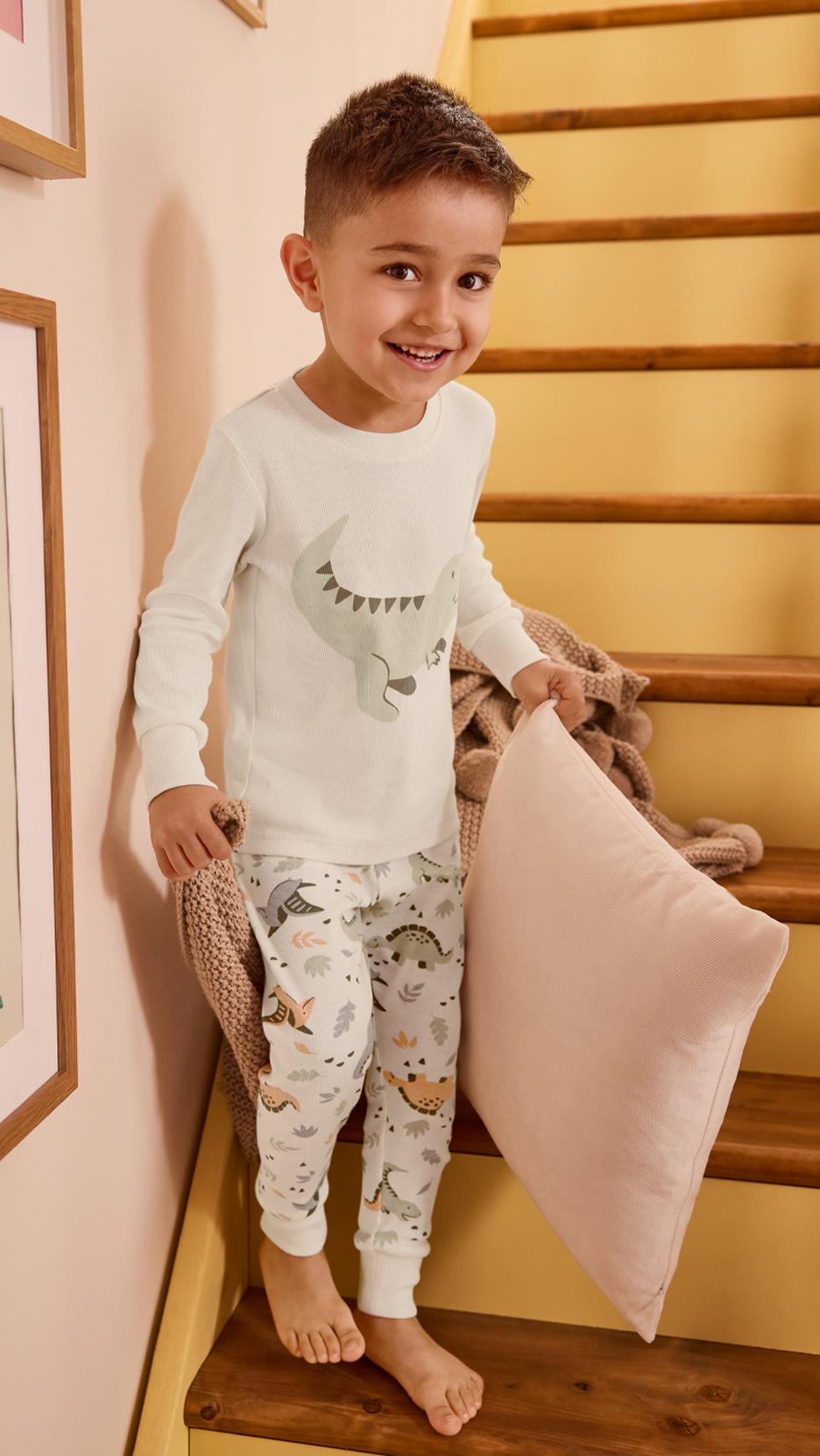Fiú dinoszaurusz mintás pizsamában, takaróval és párnával a lépcsőn.