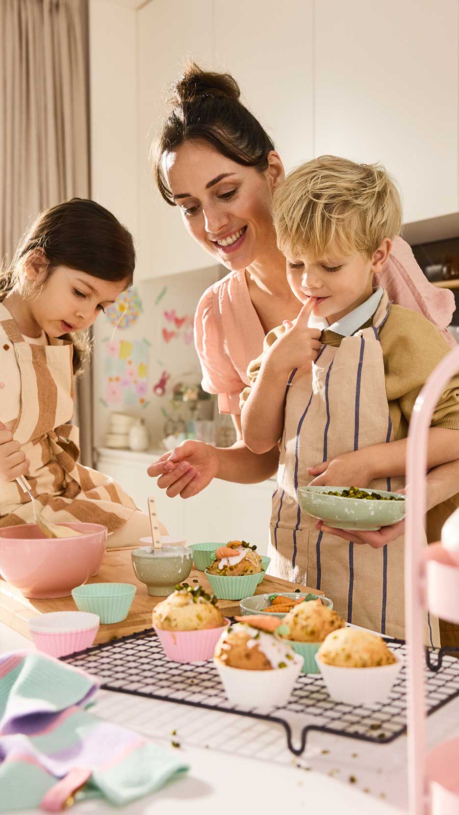 Anya és két gyermeke muffint díszít a konyhában, színes tálakkal és sütőformákkal.