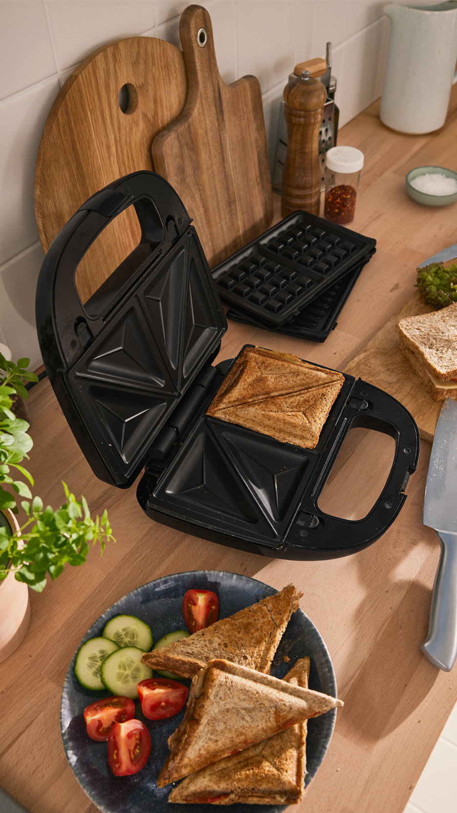 Szendvicssütő egy elkészült szendviccsel, vágódeszkák és fűszerek egy konyhapulton.