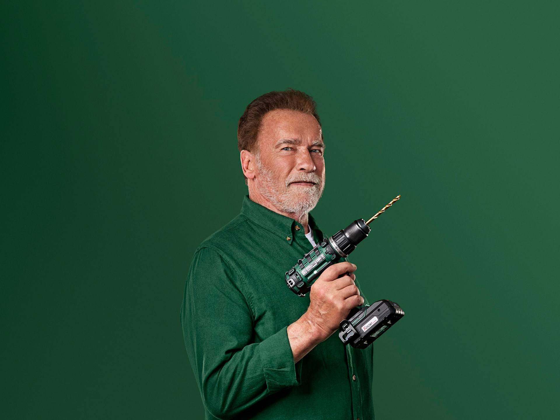 Arnold Schwarzenegger egy zöld elektromos fúrót tart zöld háttér előtt.
