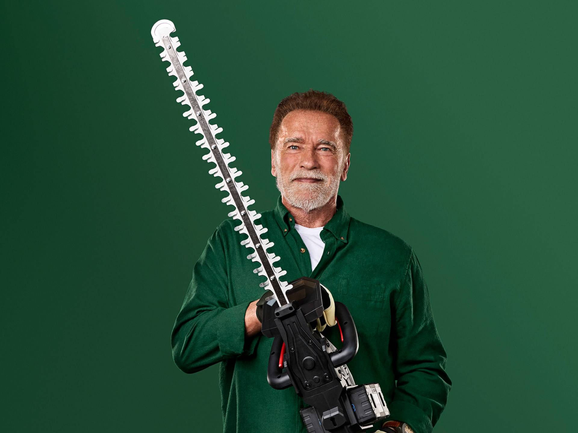 Arnold Schwarzenegger egy nagy sövényvágót tart zöld háttér előtt.