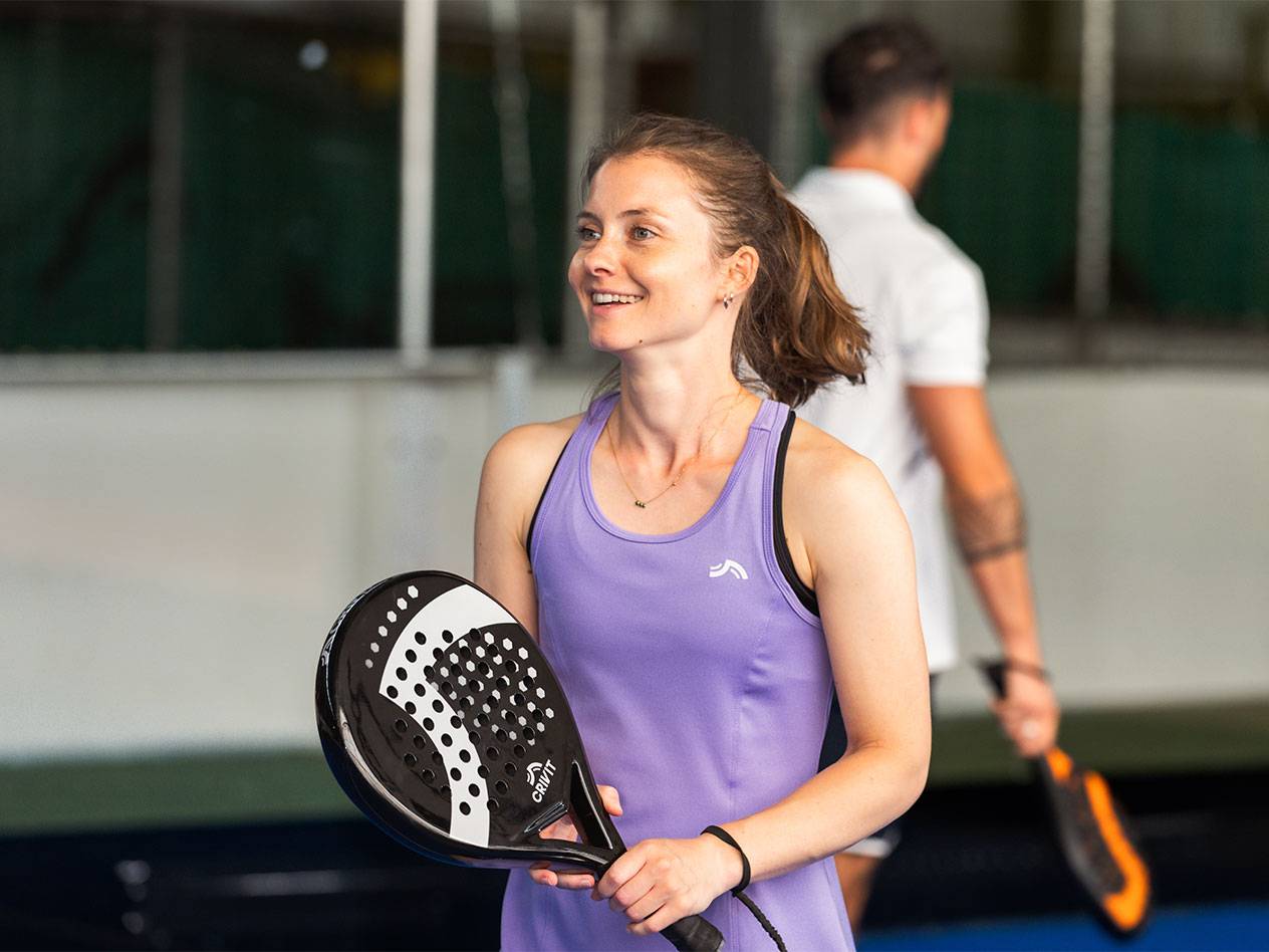 Nő lila sportruhában padelütőt tart, férfi a háttérben.