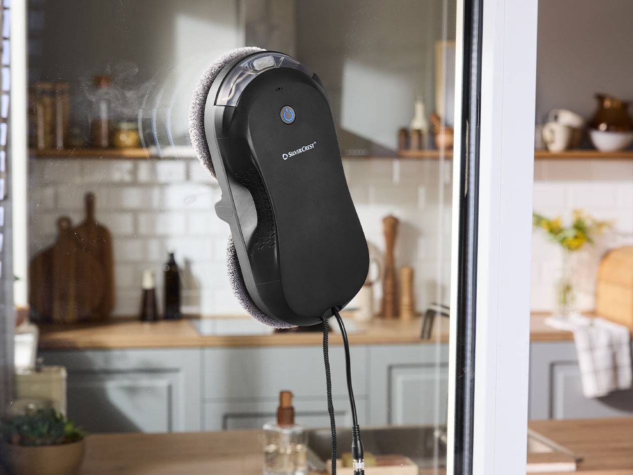 Silvercrest ablaktisztító robot egy ablaküvegen, háttérben egy konyhával.