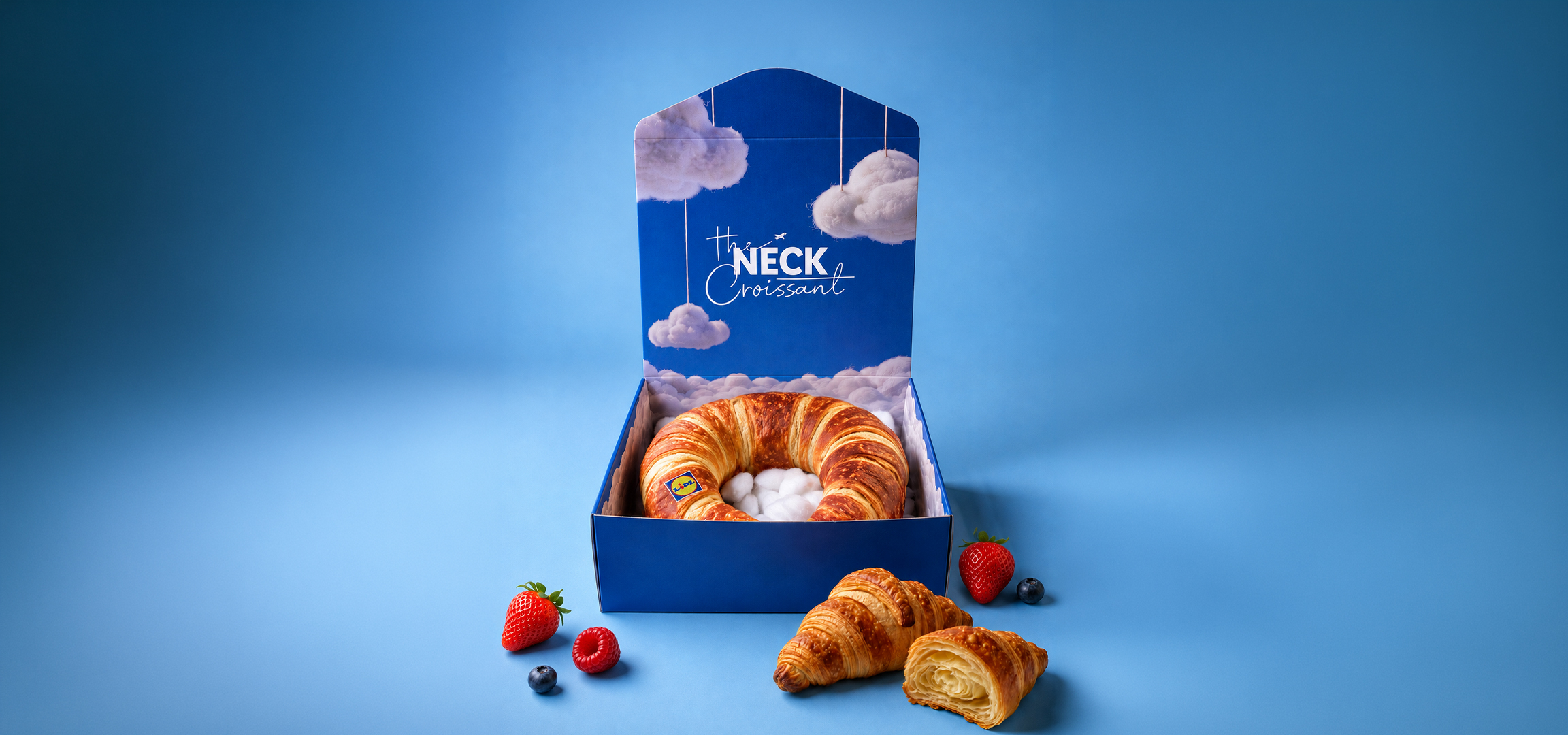 MI/AI segítségével létrehozott/módosított kép: Nyakpárna alakú croissant kék dobozban, gyümölcsökkel és péksüteményekkel körülvéve.