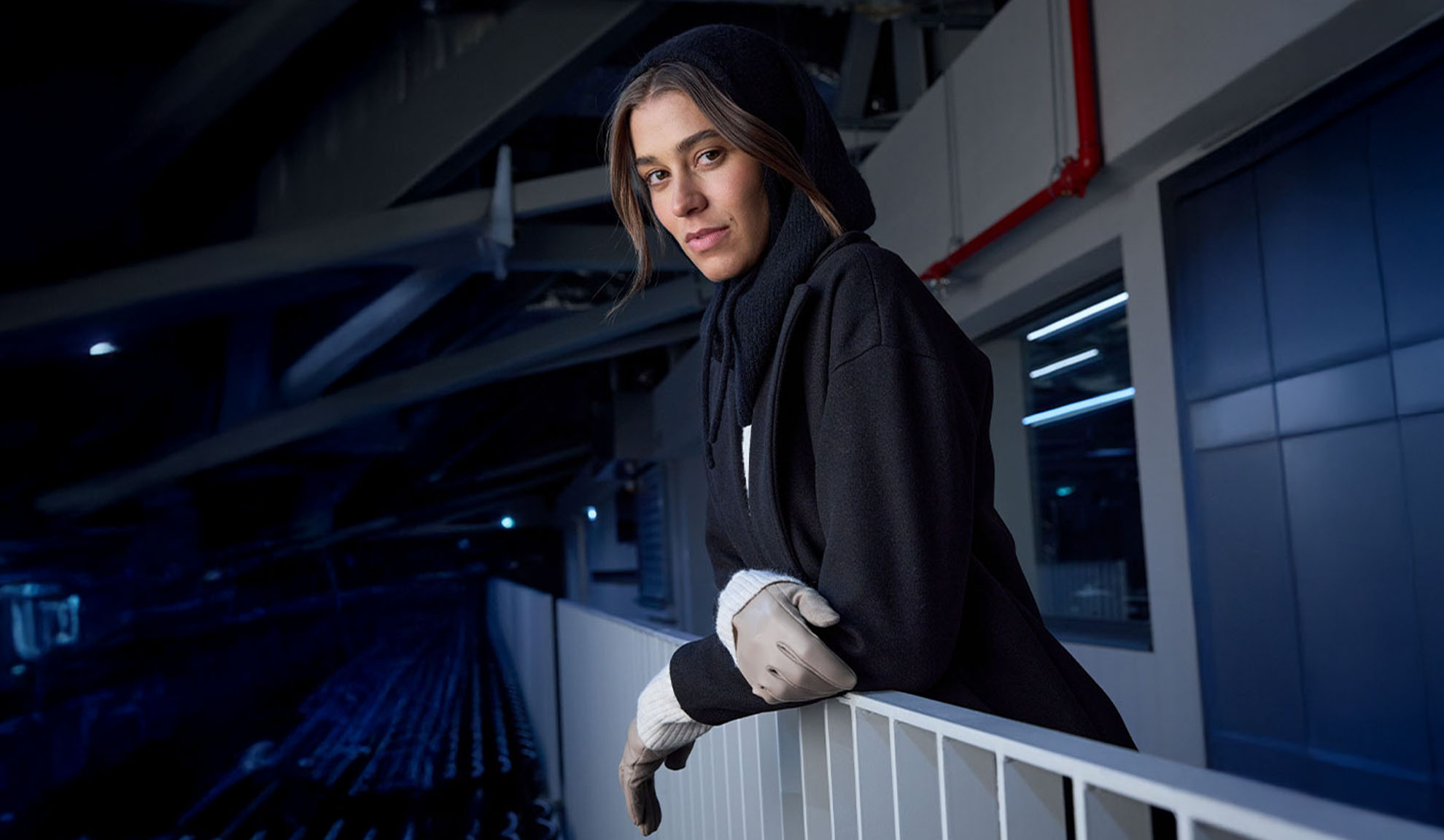 Nő fekete kabátban és kapucnis sálban, korlátra támaszkodva modern környezetben.