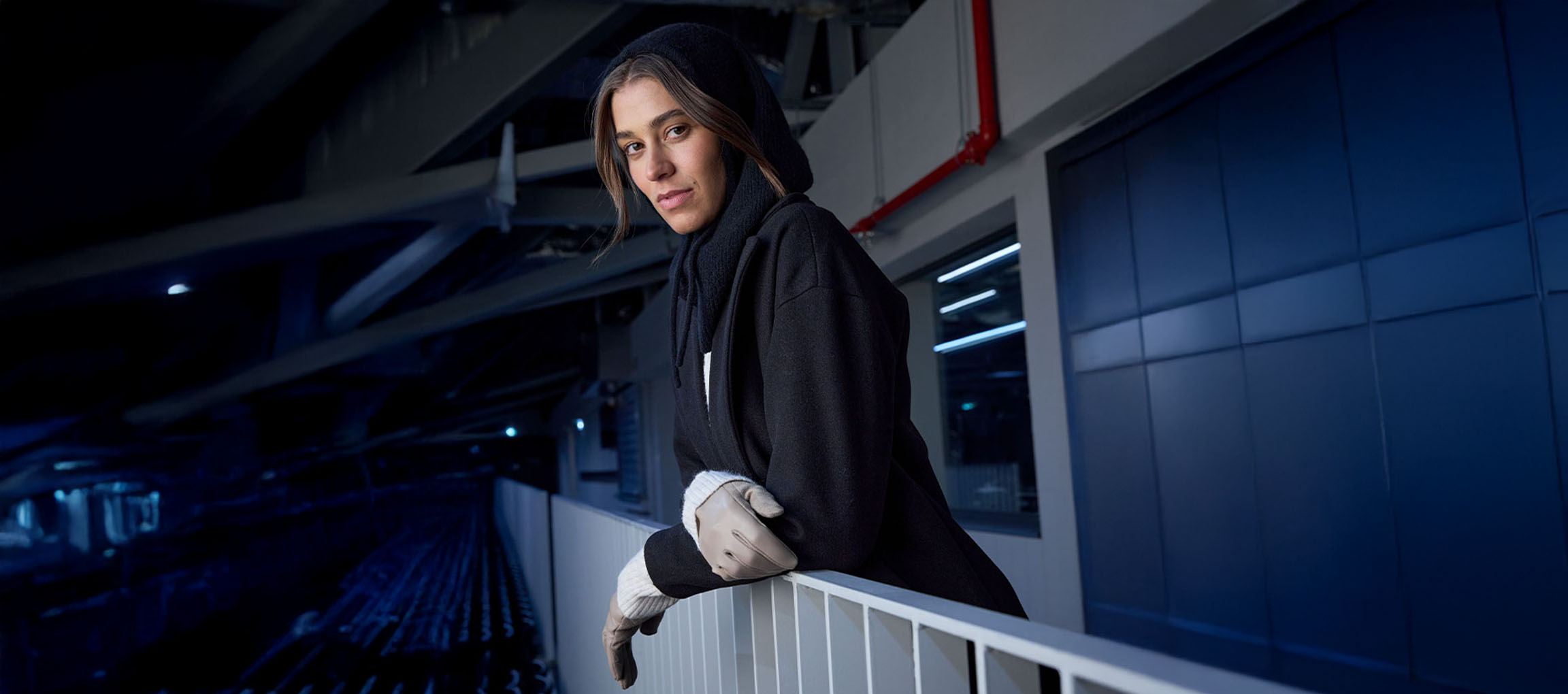 Nő fekete kabátban és kesztyűben korlátra támaszkodva városi környezetben.