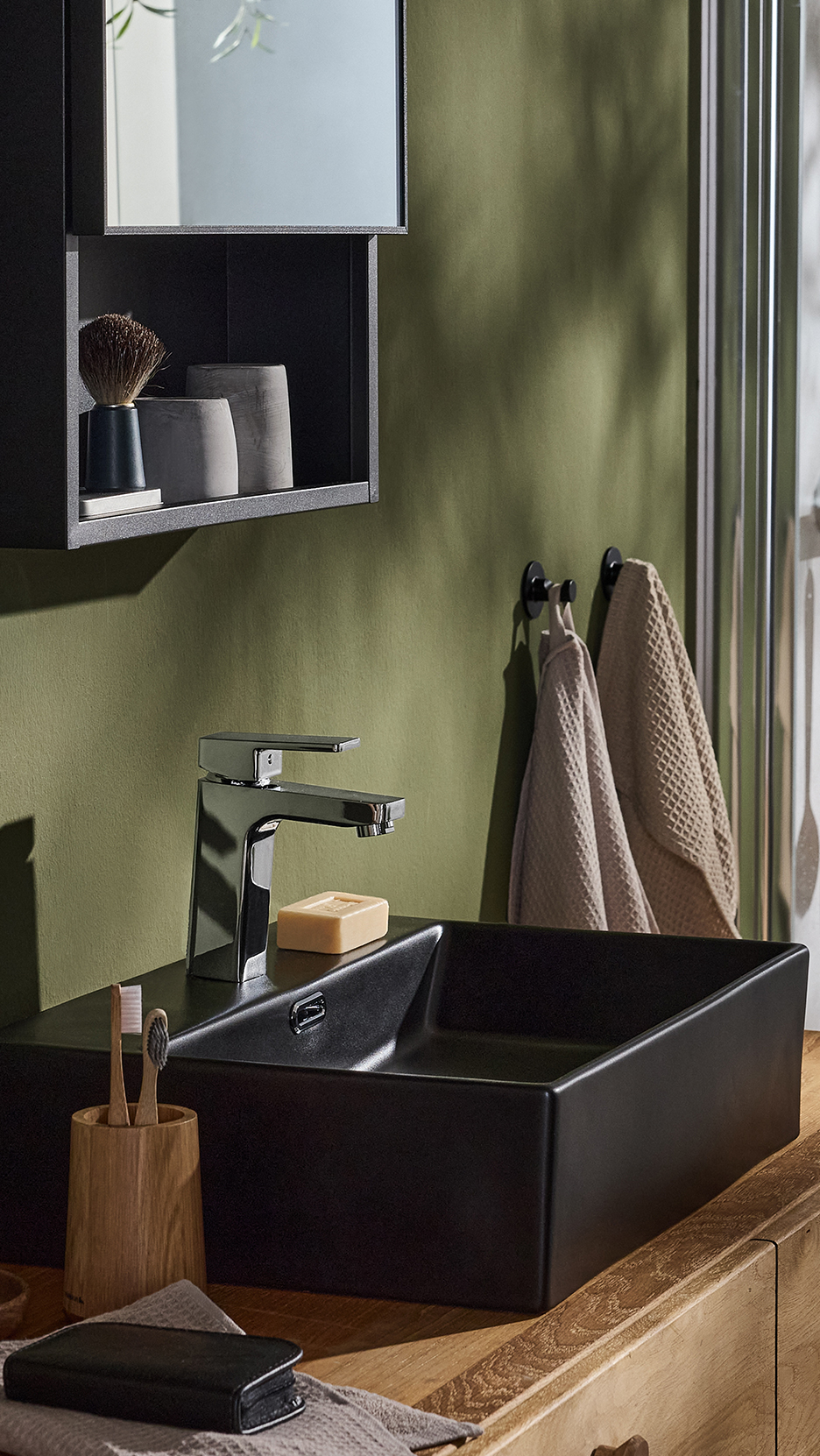 Fekete mosdókagyló króm csapteleppel, zöld fal előtt, fa fürdőszobabútoron.