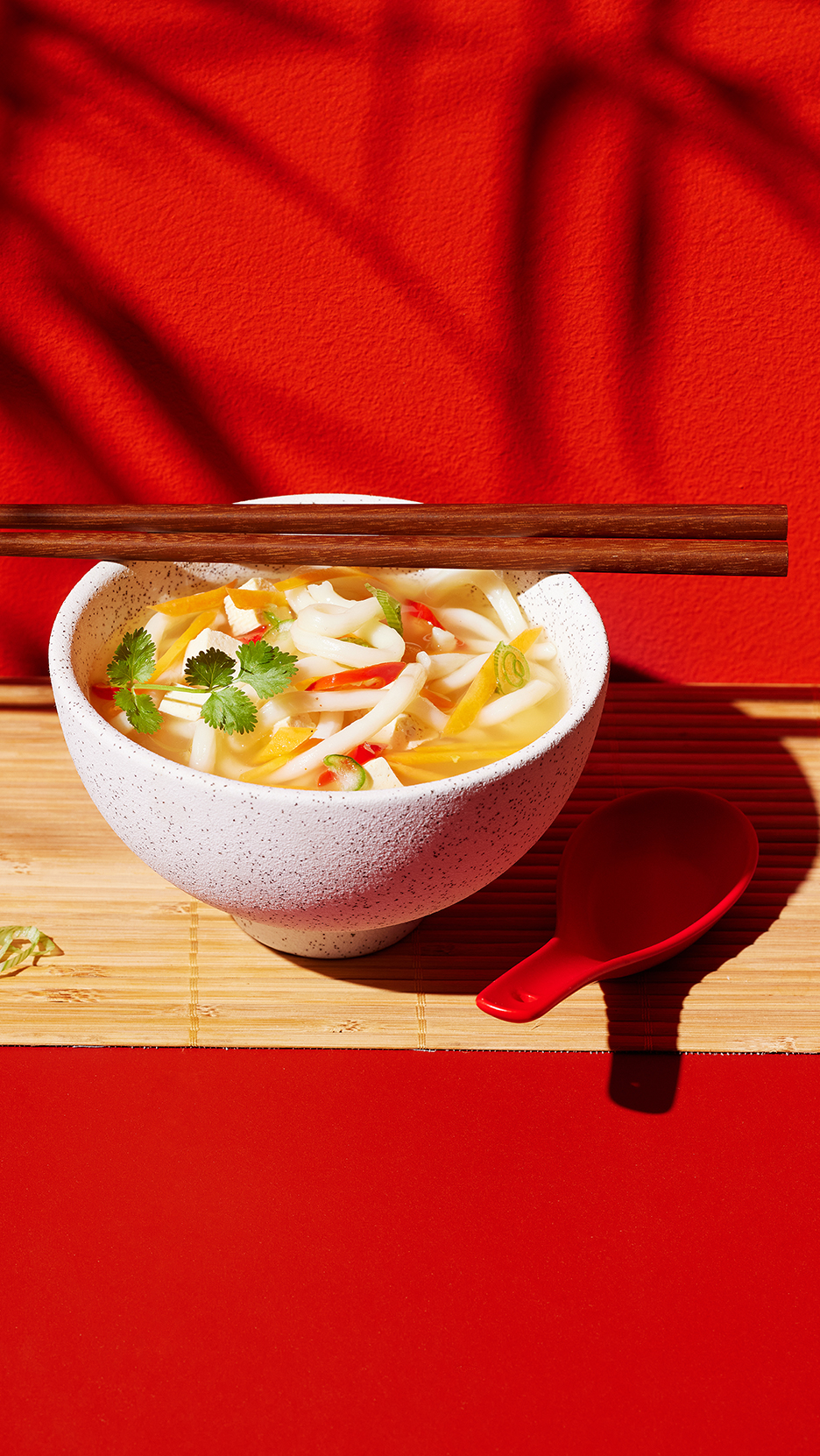 Tésztaétel tofuval, zöldségekkel és korianderrel, pálcikákkal és kanállal, piros háttér előtt.