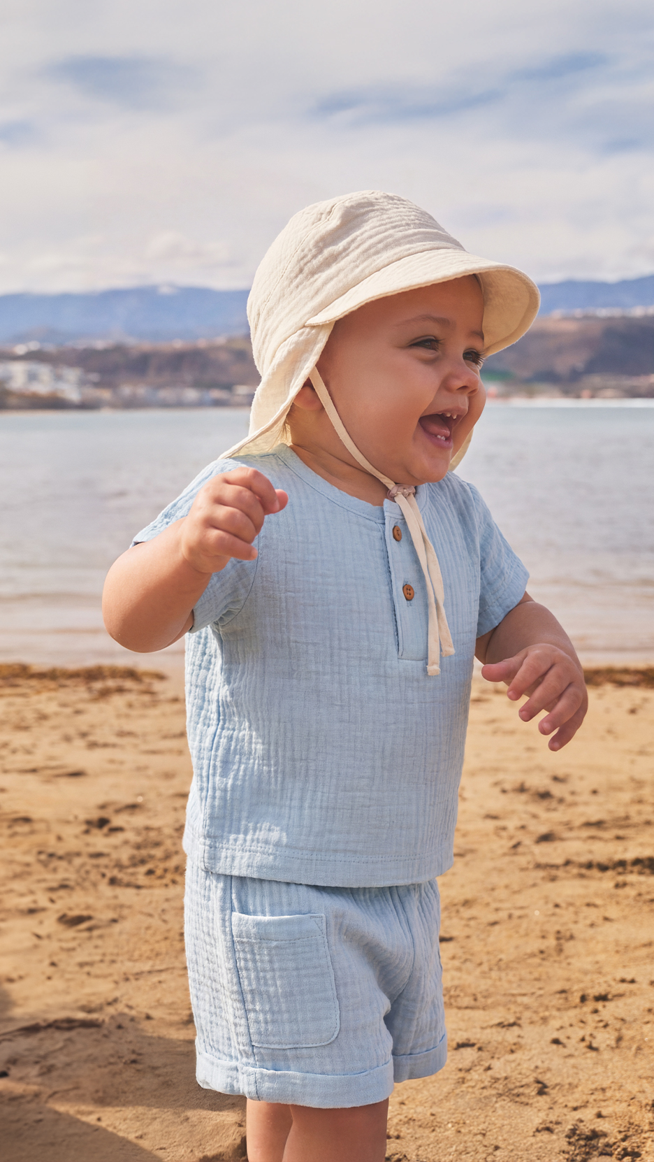 Kisfiú világoskék muszlin szettben és bézs kalapban a tengerparton.