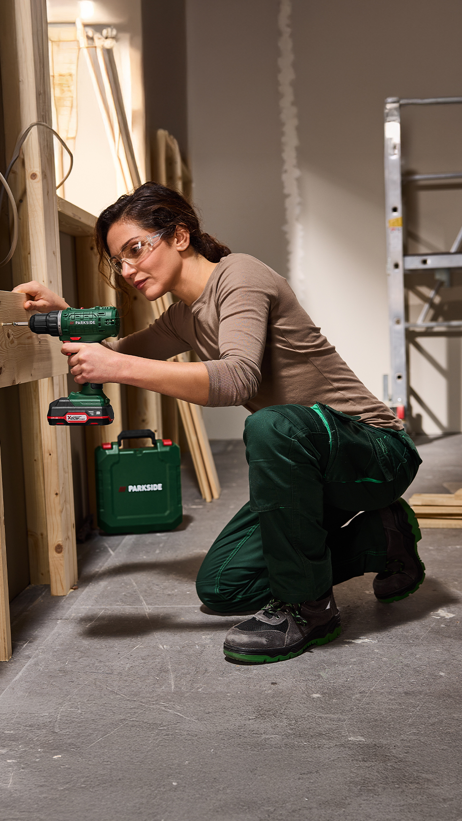Nő akkus fúróval dolgozik egy építkezésen, zöld szerszámosládával a földön.