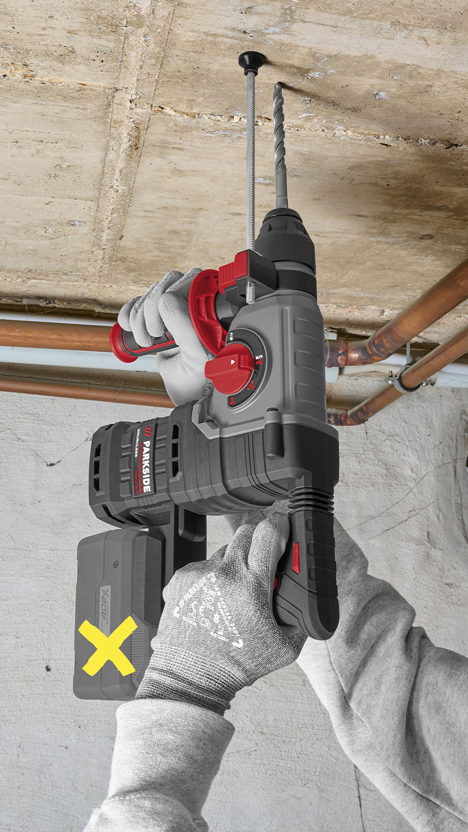 Fúrókalapácsot tartó kezek, mennyezetbe fúrás közben, rézcsövekkel a háttérben.