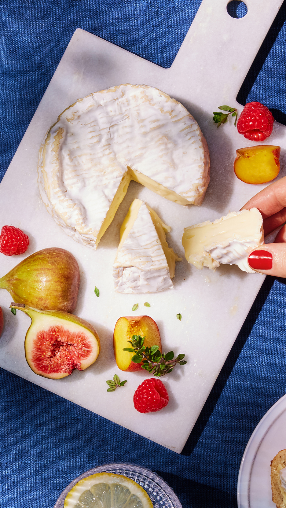 Márványtálon camembert sajt, füge, málna és őszibarack szeletek, egy kéz egy darab sajtot tart.