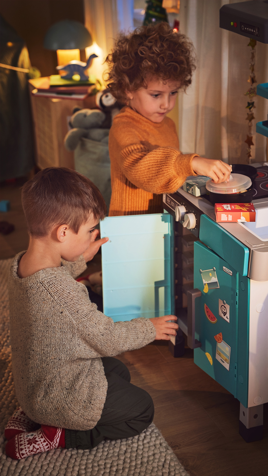Két gyermek játszik egy játék konyhával, az egyik a sütő ajtaját nyitja, a másik a főzőlapon főz.