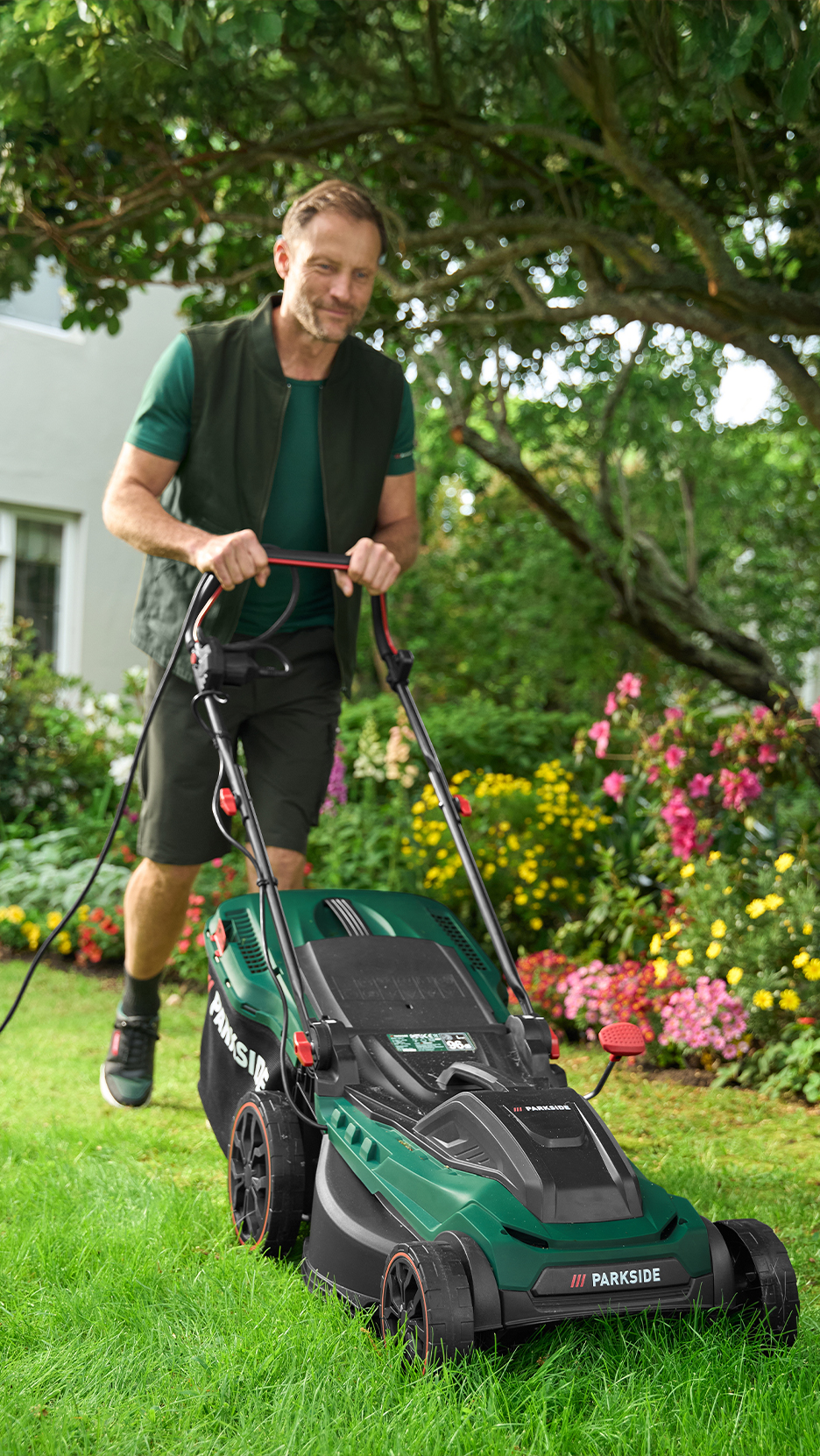 Férfi zöld fűnyíróval nyírja a füvet egy virágos kertben.