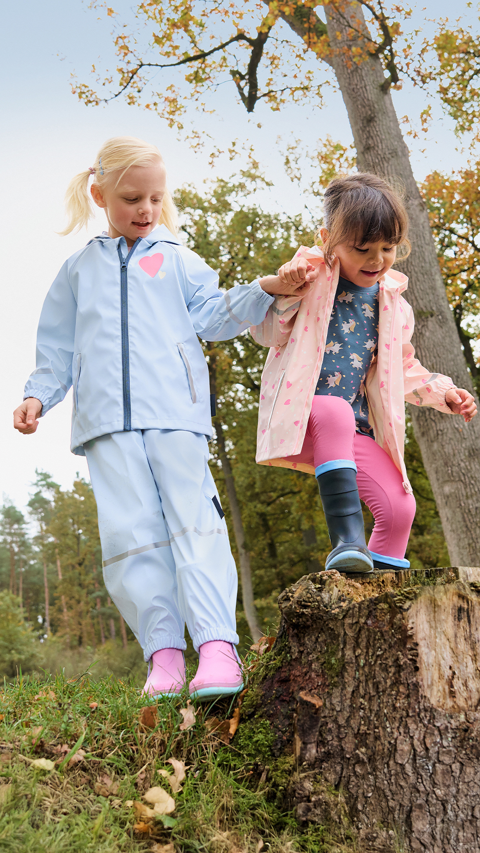 Két kislány esőruhában és gumicsizmában játszik az erdőben.
