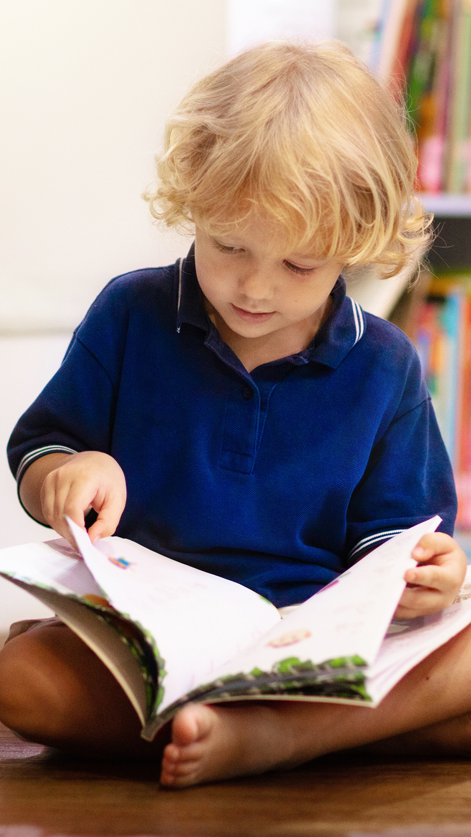 Egy szőke hajú gyermek kék pólóban olvas egy könyvet a földön ülve.