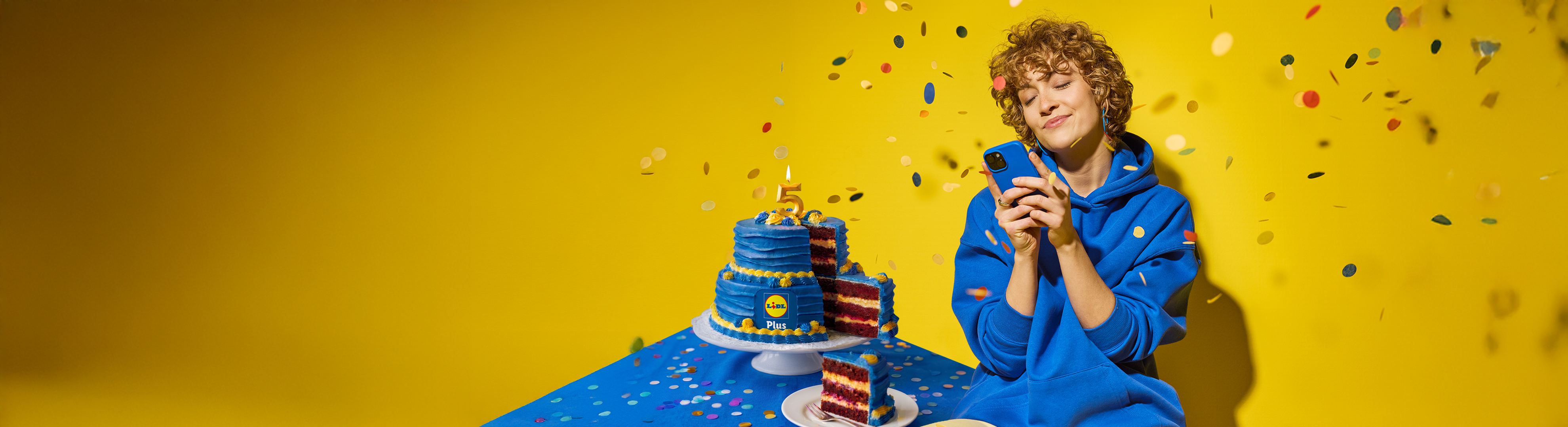 Kék torta 5-ös gyertyával és egy nő telefonnal, konfetti esik rájuk.