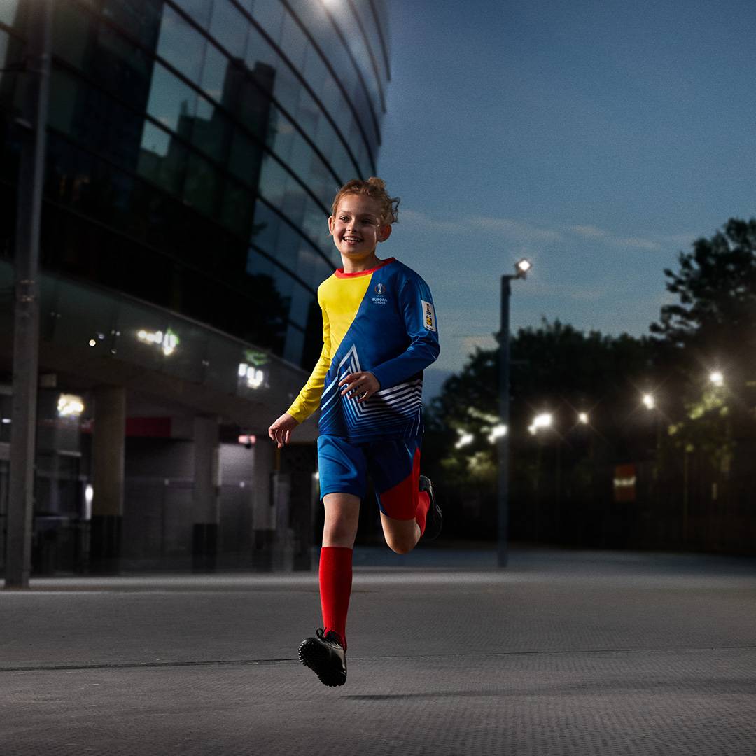 Fiú futballmezben és rövidnadrágban fut egy stadion előtt, este.