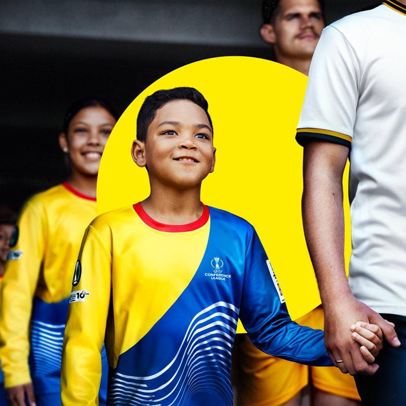 Fiatal fiú sárga és kék focimezben egy felnőtt kezét fogja.