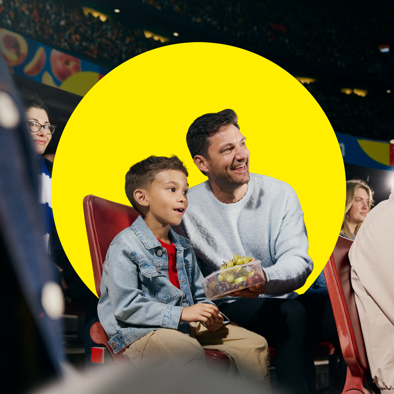 Apa és fia gyümölcsöt fogyasztanak egy dobozból sporteseményen.