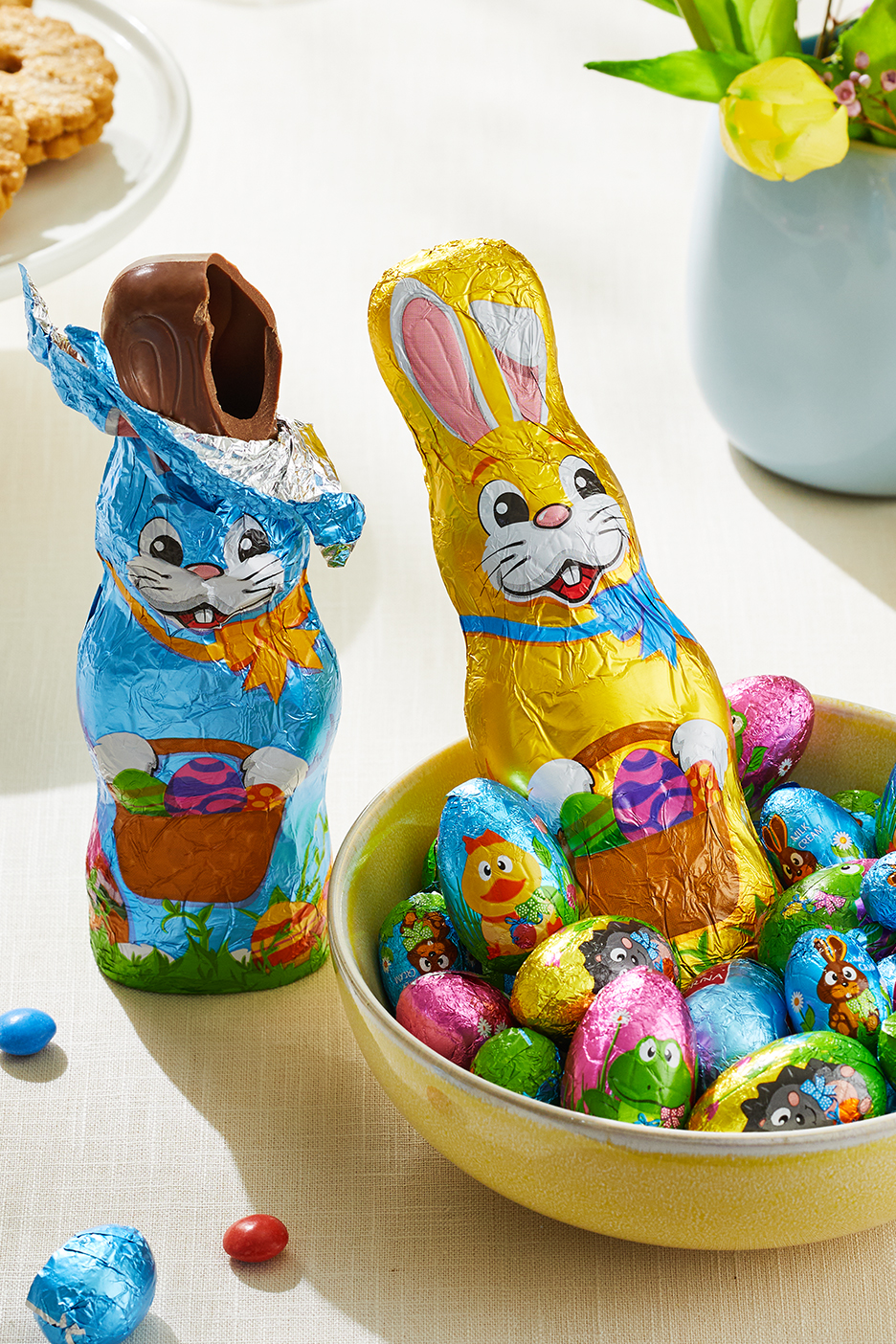 Húsvéti csokoládé nyuszik és tojások egy tálban, kekszekkel és virágokkal.