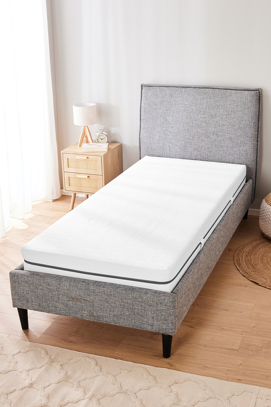 Szürke szövet ágykeret fehér matraccal, éjjeli szekrénnyel és lámpával egy hálószobában.