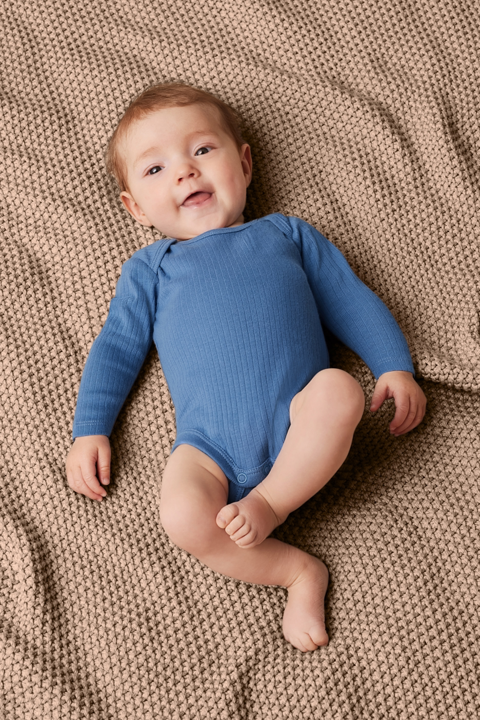 Kék színű, hosszú ujjú bodyt viselő baba fekszik egy bézs színű takarón.