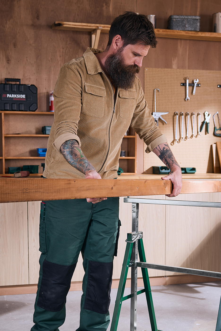 Férfi Parkside szerszámosládával és szerszámokkal egy műhelyben, fát tartva egy fűrészbakon.