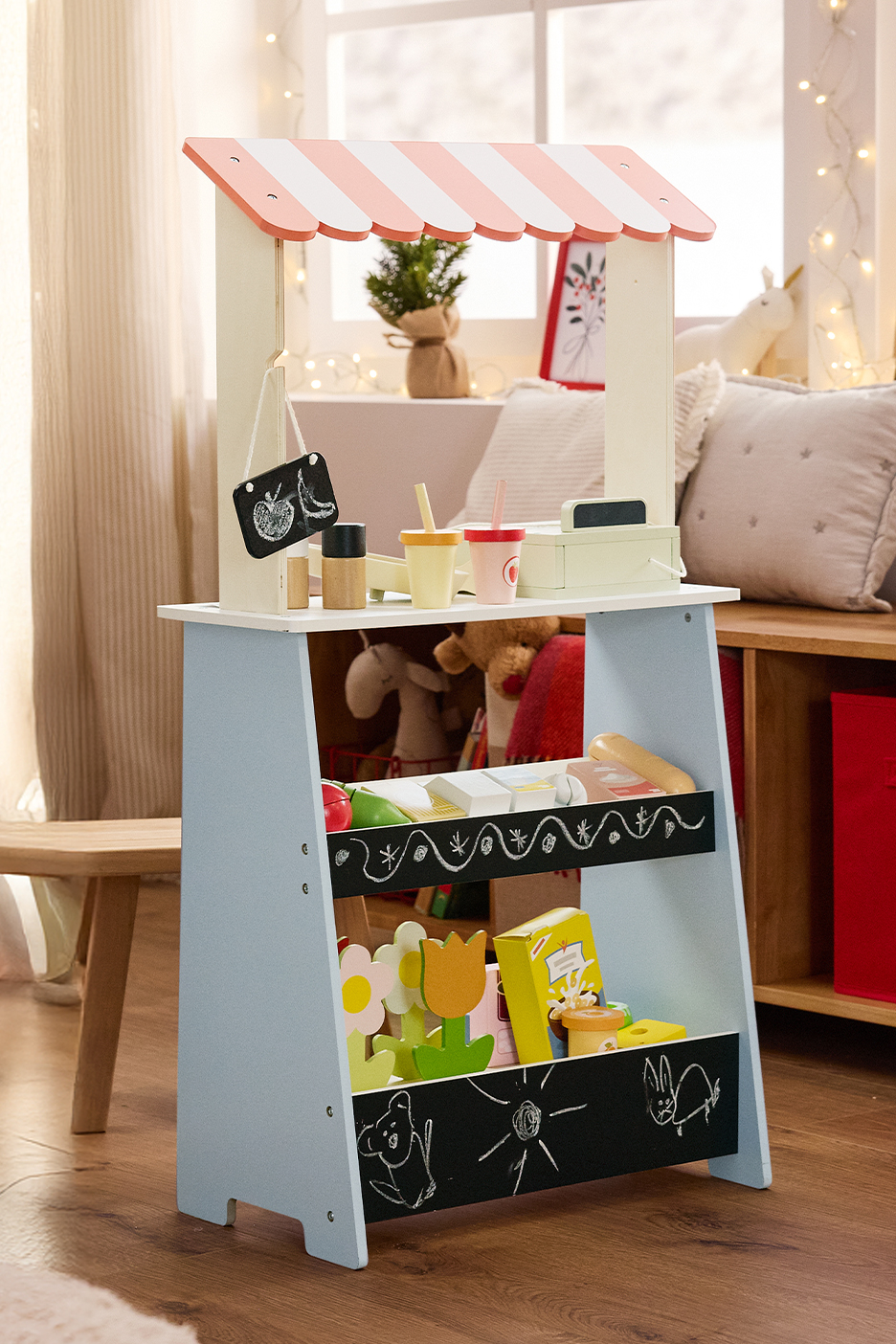 Játék stand rózsaszín-fehér csíkos tetővel, krétatáblával és játék élelmiszerekkel egy gyerekszobában.