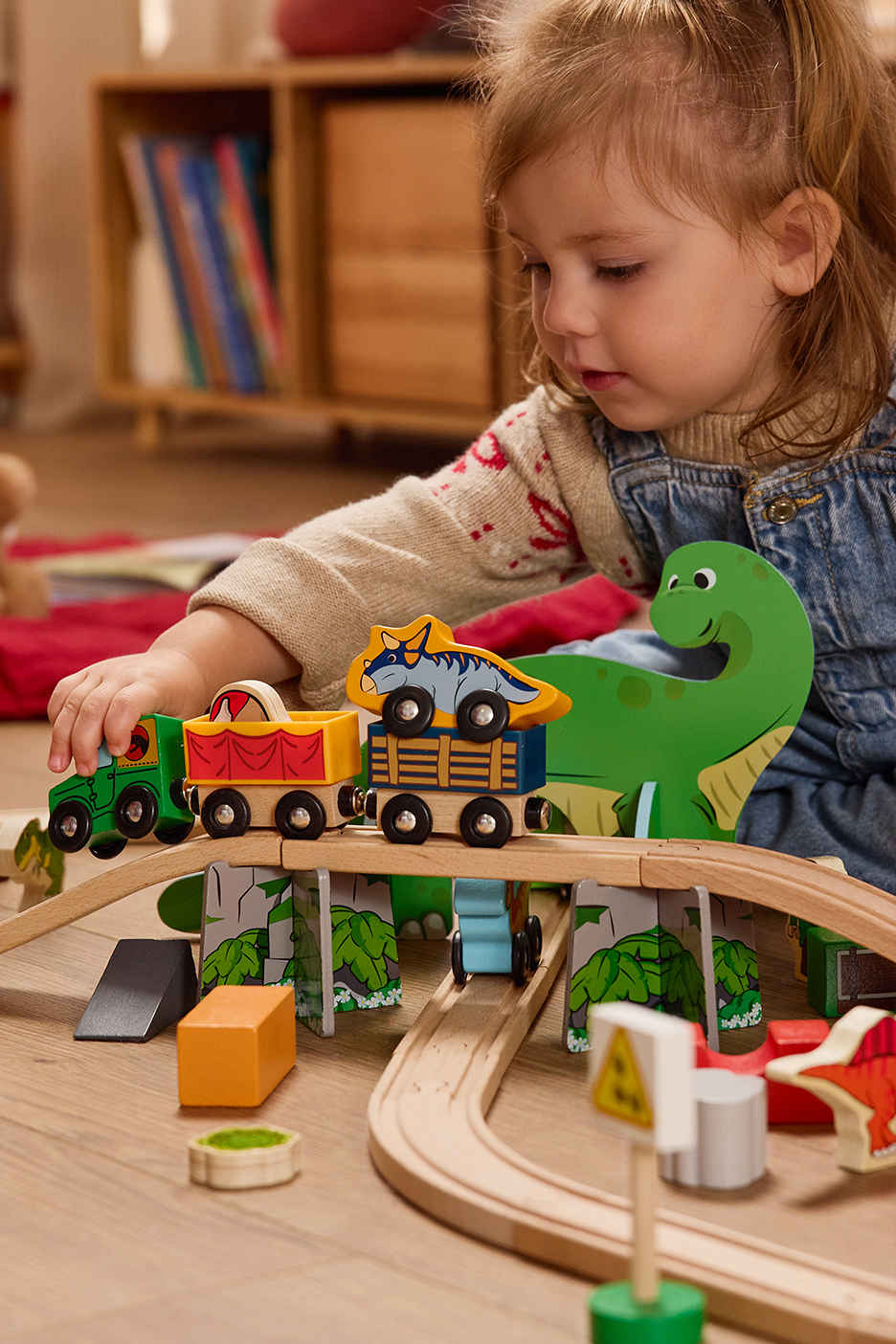 Kislány játszik egy dinoszaurusz témájú fa vonatkészlettel a padlón.