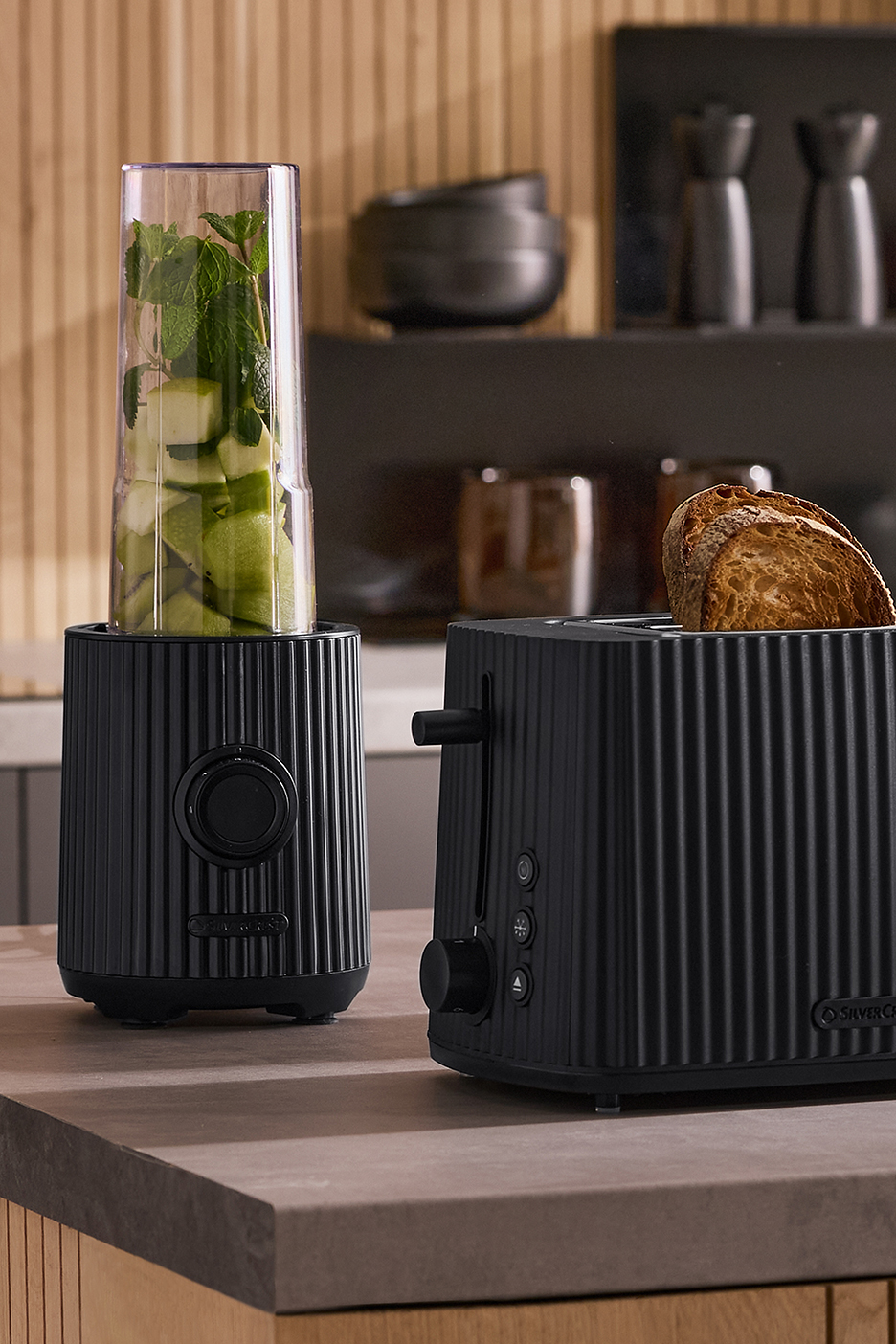 Fekete bordázott turmixgép zöld almával és mentával, valamint egy kenyérpirító pirítóssal egy konyhapulton.