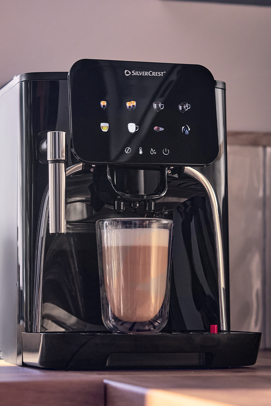 Fekete automata kávéfőző tejhabos kávéval egy üvegpohárban.