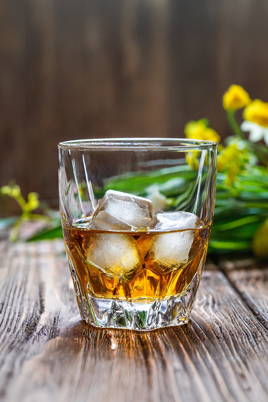 Whisky jéggel egy pohárban, fa asztalon, virágokkal a háttérben.