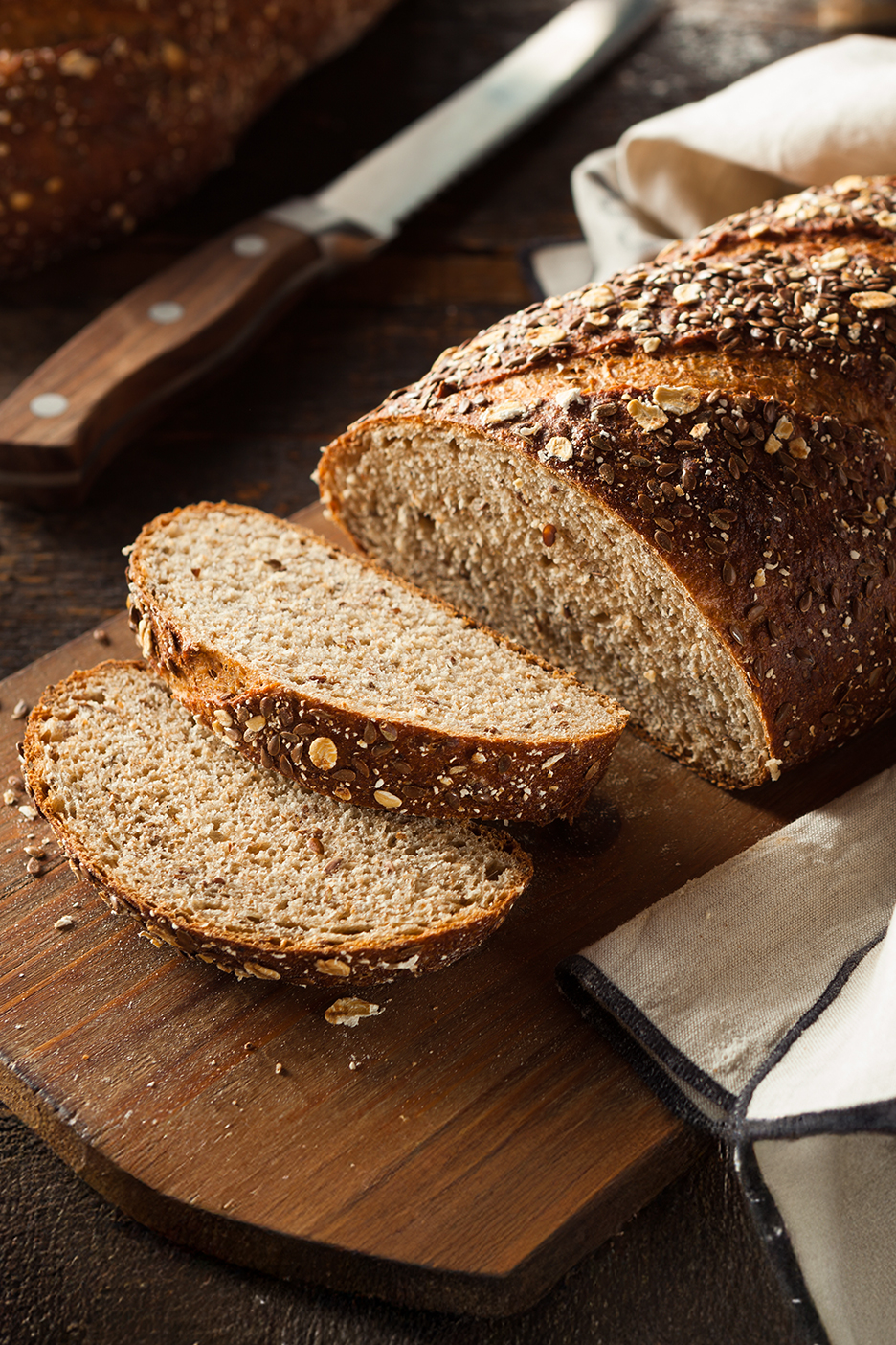 Szeletelt magvas kenyér vágódeszkán, késsel és konyharuhával.