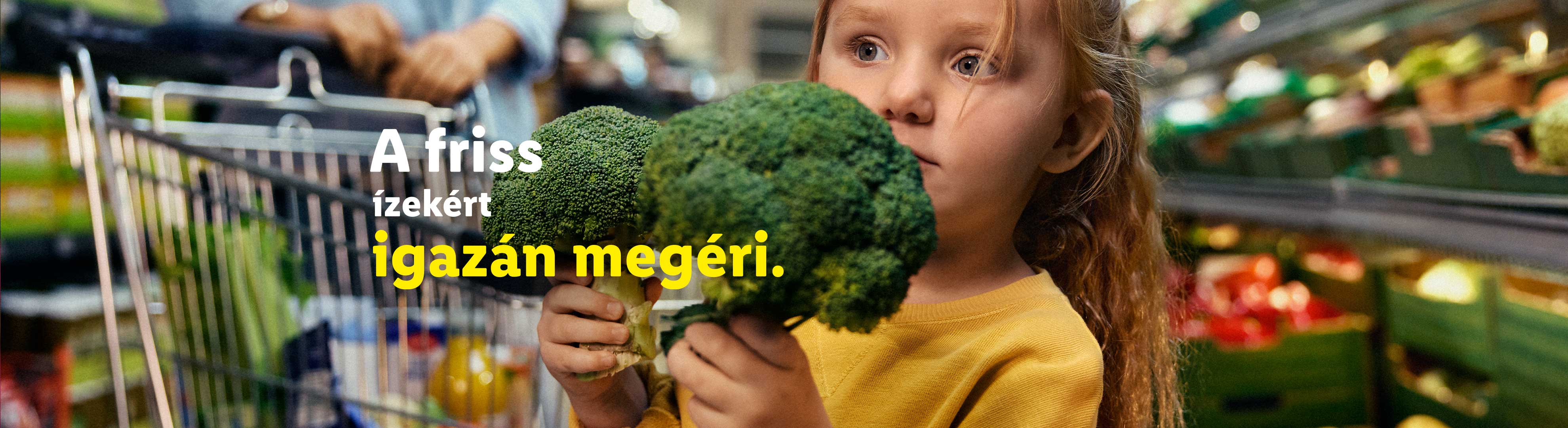 Egy kislány két brokkolit tart egy szupermarketben, a háttérben egy bevásárlókocsi látható.