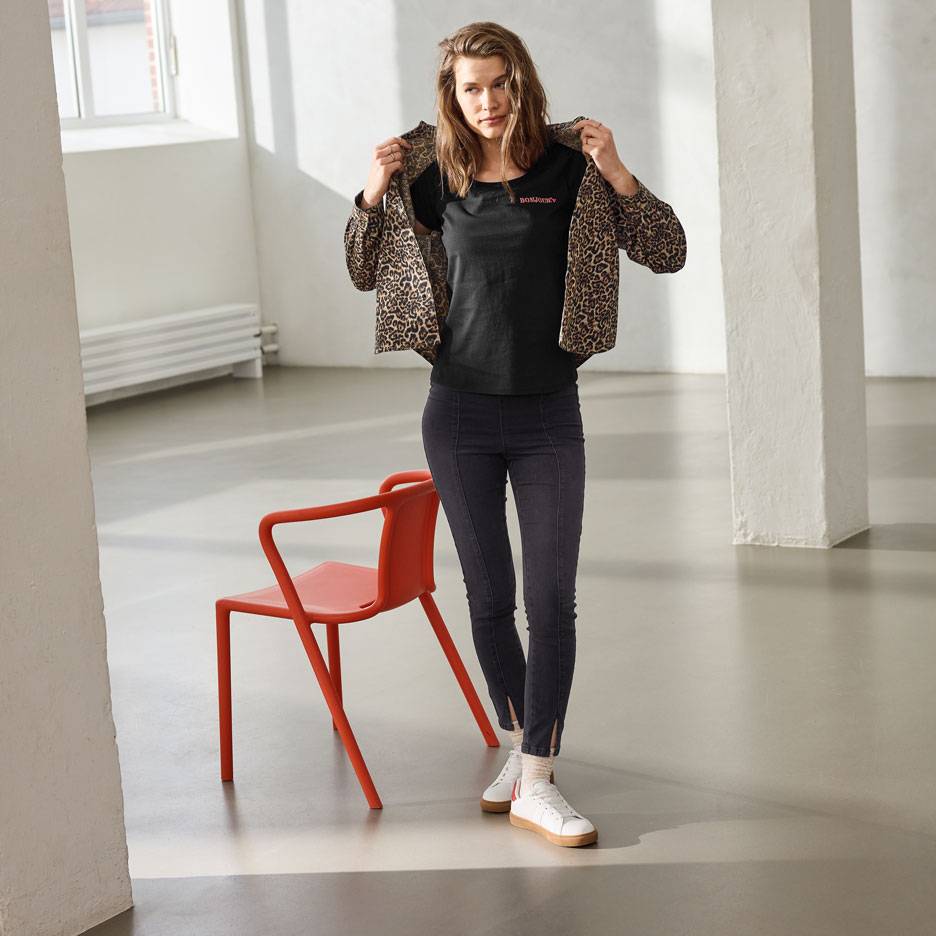 Nő fekete feliratos pólóban, farmerben és leopárdmintás kabátban, piros szék mellett állva.