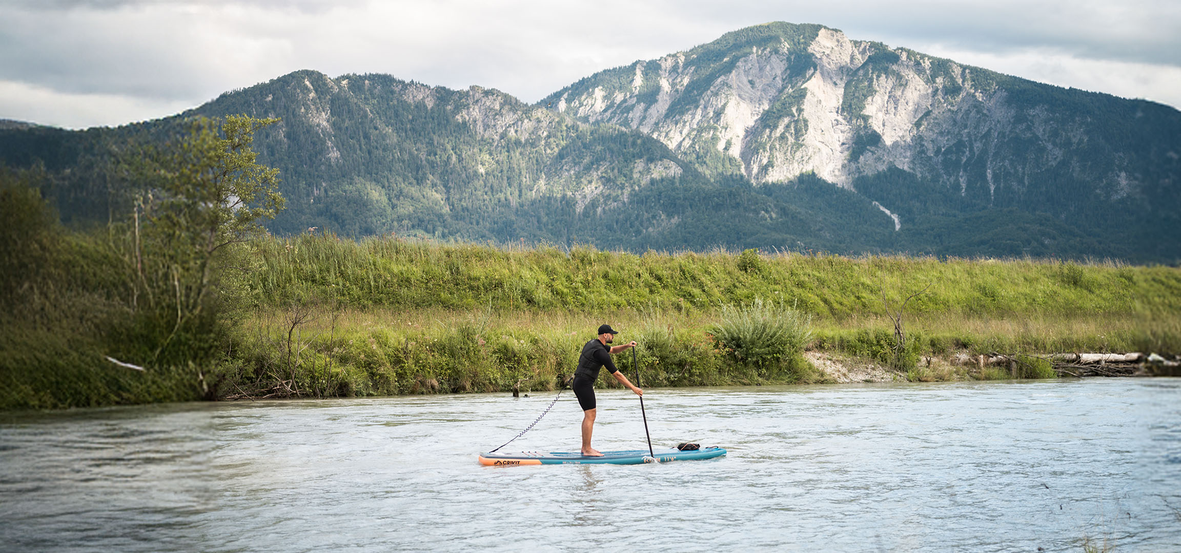 Férfi paddleboardozik egy folyón, hegyekkel és buja növényzettel a háttérben.